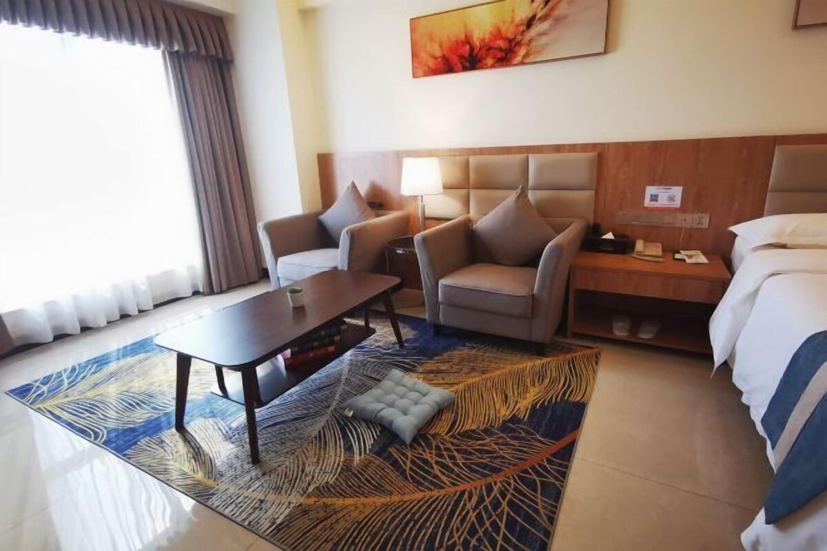A hotel room with a bed, a sofa, a table, and a chair. There is a window with curtains, a painting on the wall, and a rug on the floor.
