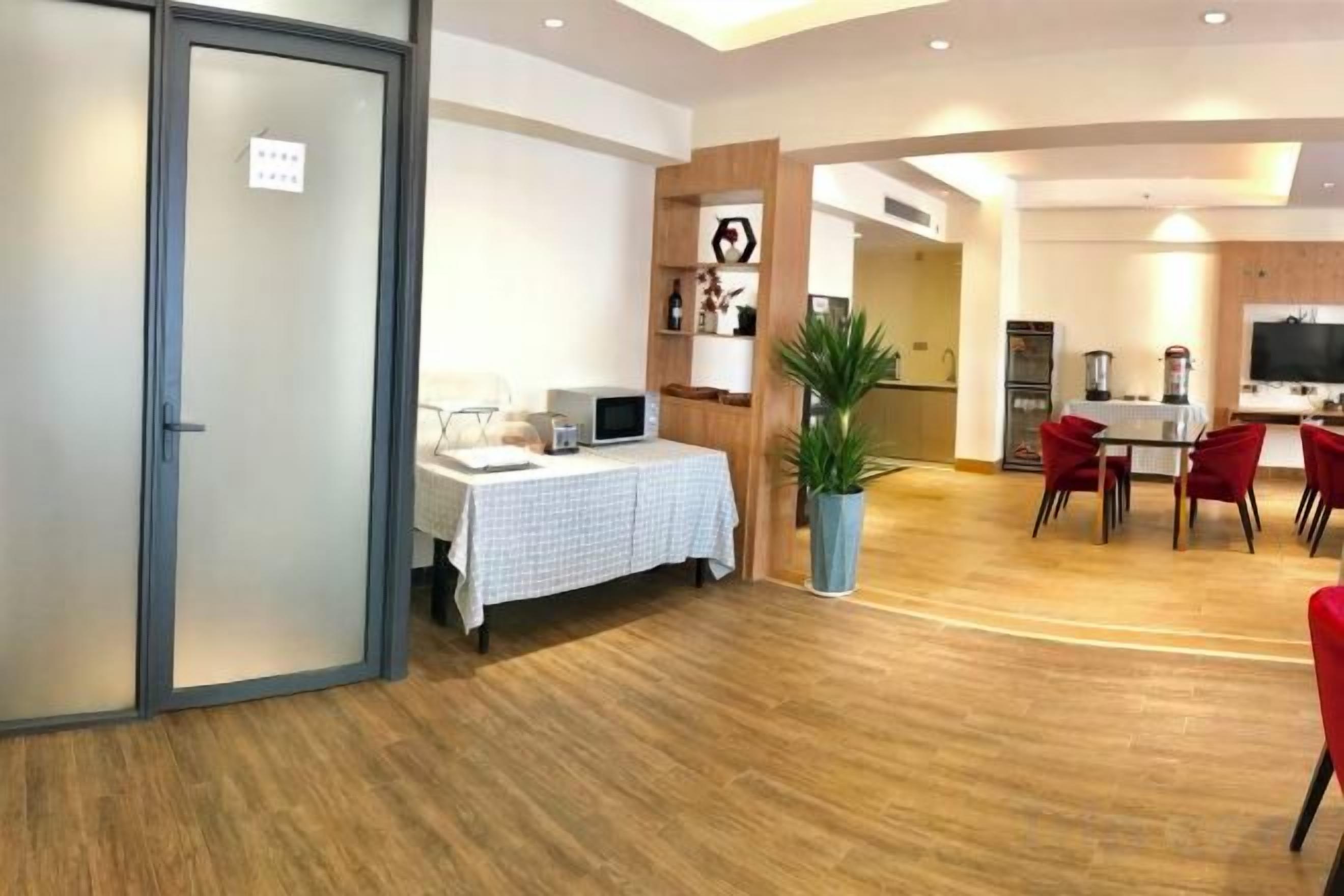 Habitación de hotel con cama, mesita de noche y una planta grande en maceta. Cuenta con un comedor con mesa y sillas, y un televisor montado en la pared.