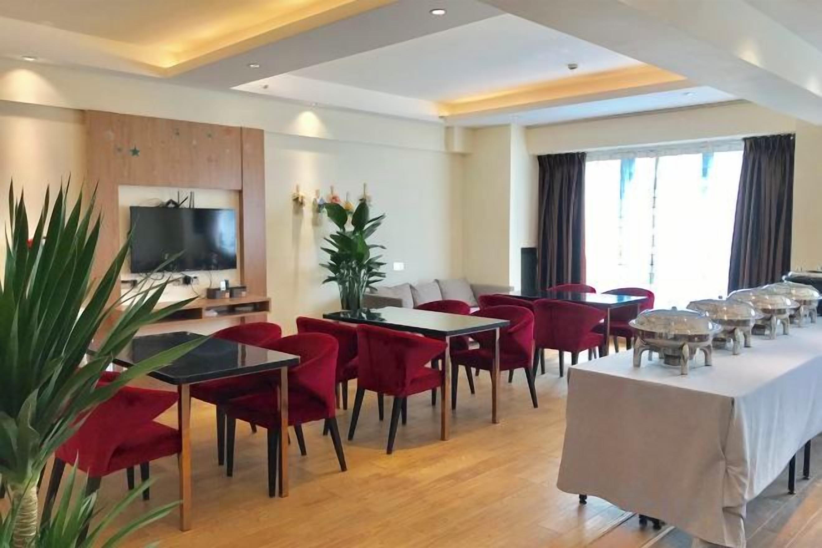 Comedor de hotel con sillas rojas, una mesa larga, un televisor y un ventanal grande.