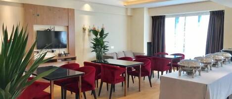 Comedor de hotel con sillas rojas, una mesa larga, un televisor y un ventanal grande.