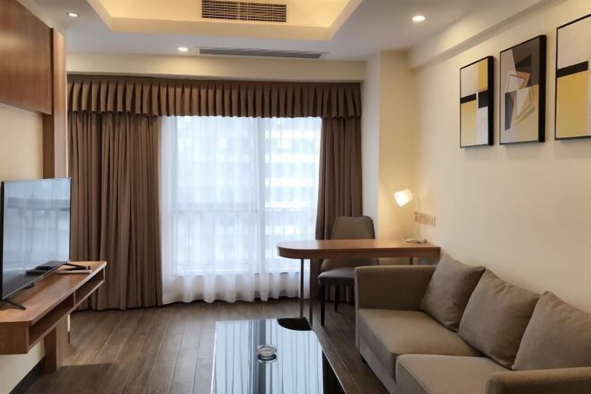 A modern hotel room with a flat-screen TV, a wooden coffee table, a sofa, a dining table with chairs, and a large window with curtains.