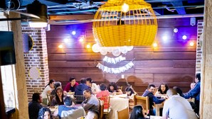 Een restaurant met houten wanden en tafels, mensen aan het dineren, en gele lampen die aan het plafond hangen.