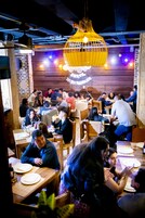 Un restaurante con paredes y mesas de madera, personas cenando y luces amarillas colgantes.