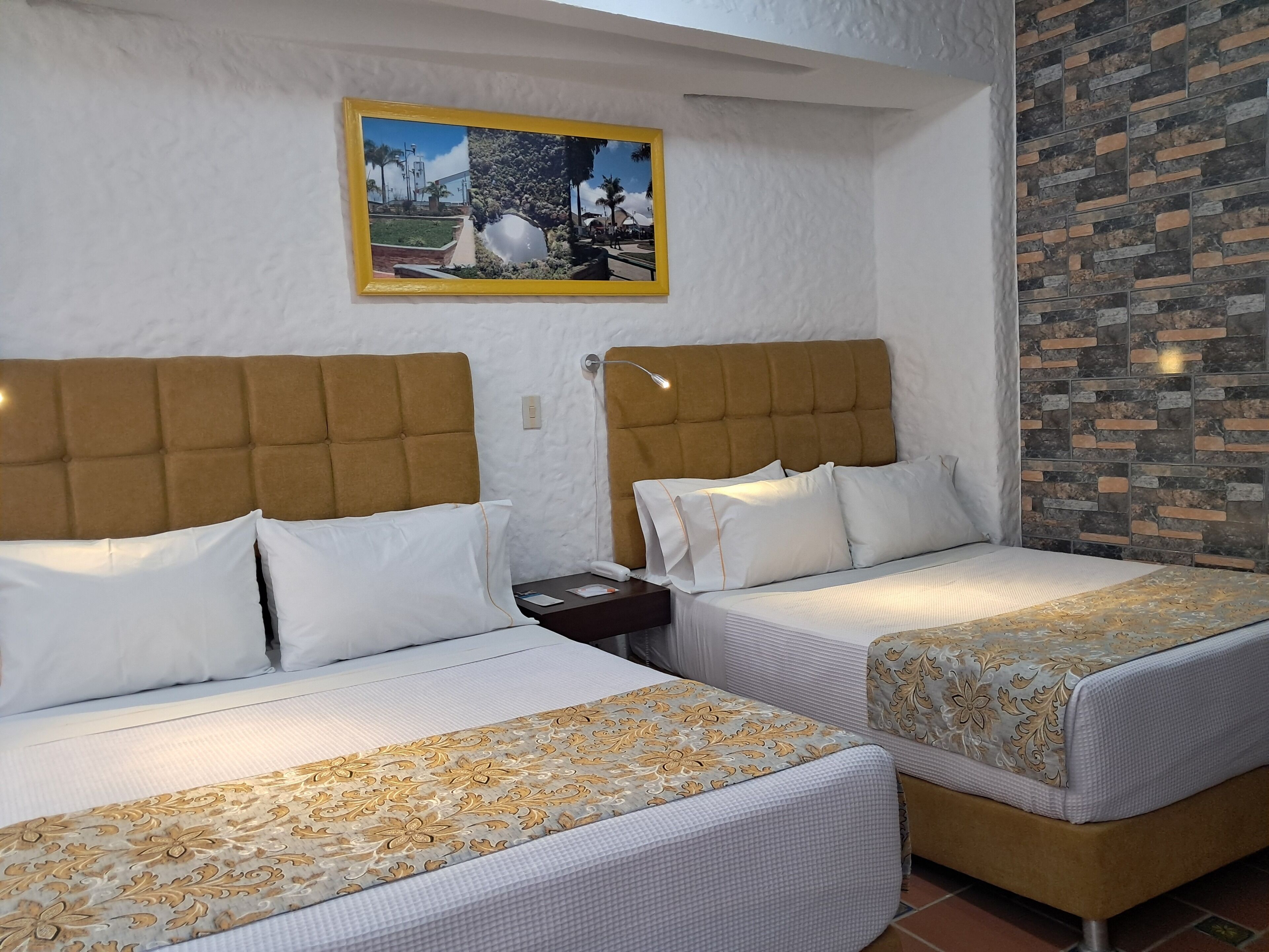Habitación de hotel con dos camas, cada una con cabecero y ropa de cama blanca. Hay un cuadro en la pared sobre las camas, y una pared con diseño de ladrillos a un lado.
