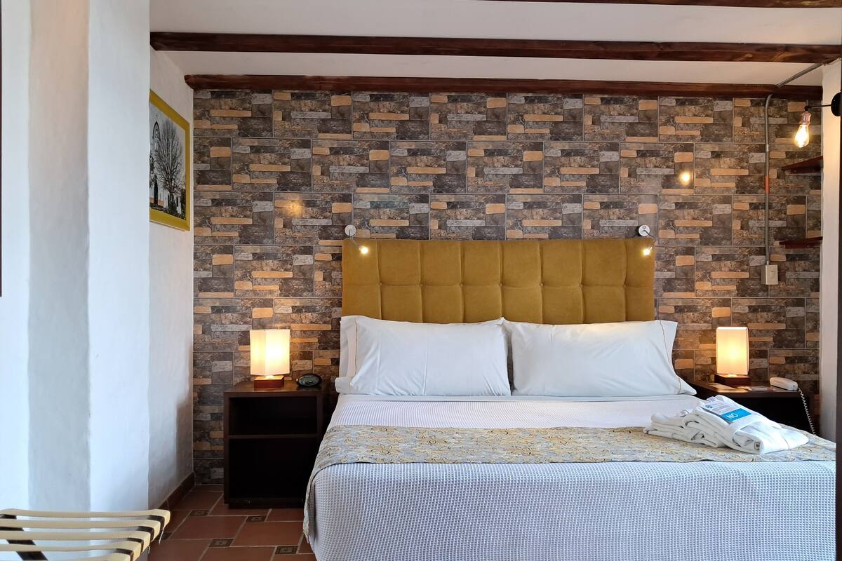 Un dormitorio con cabecera de piedra, cama con ropa blanca, dos lámparas de noche y una mesita de noche con lámpara y teléfono.