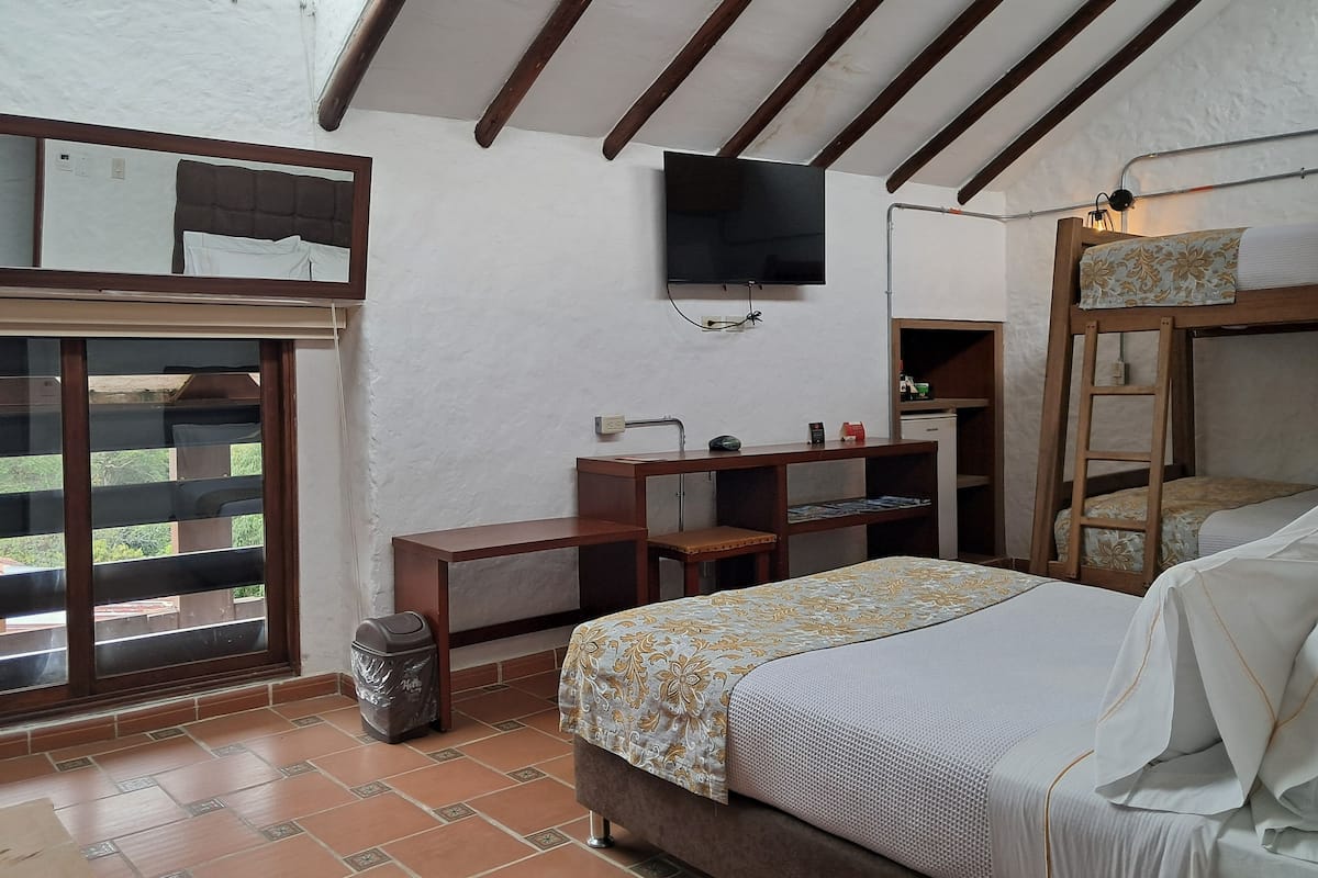 Un dormitorio con literas, un escritorio con silla, un televisor montado en la pared y una ventana con vistas a la vegetación.