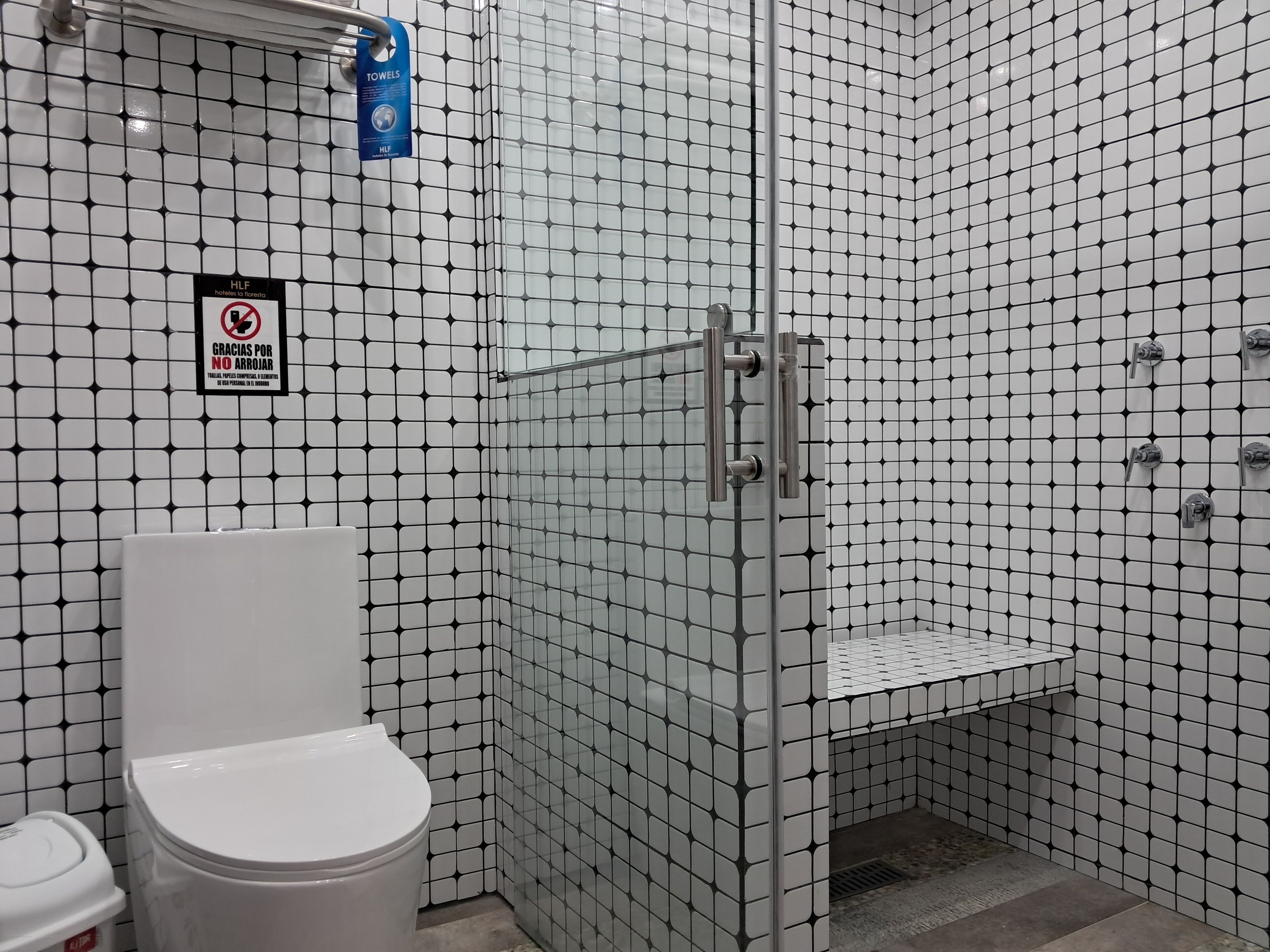 Un baño con pared de azulejos blancos, cabina de ducha con mampara de vidrio, inodoro y un cubo de basura.