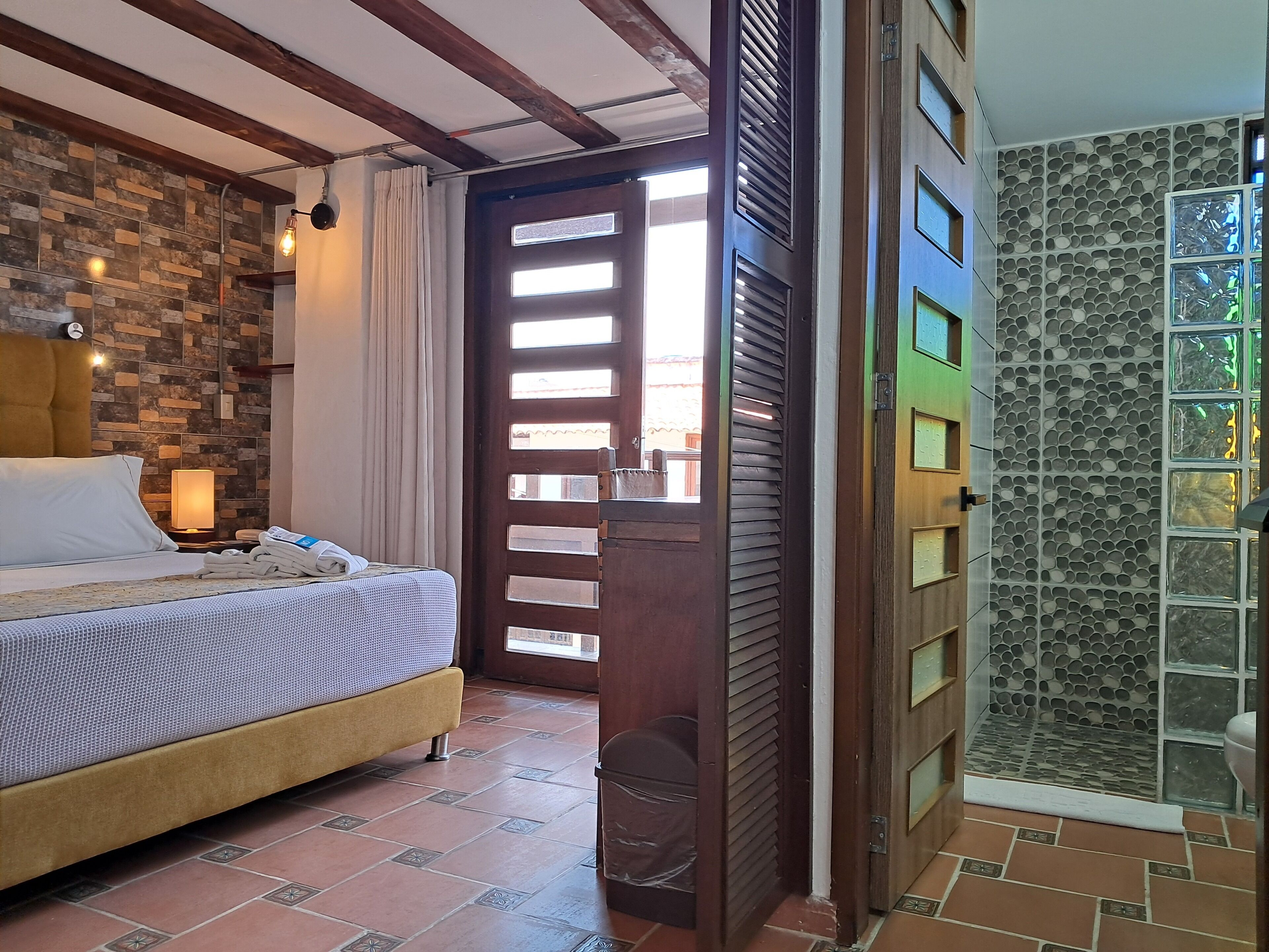 Habitación de hotel con cama, mesita de noche, lámpara, baño con puerta de vidrio y pared con diseño de piedra.