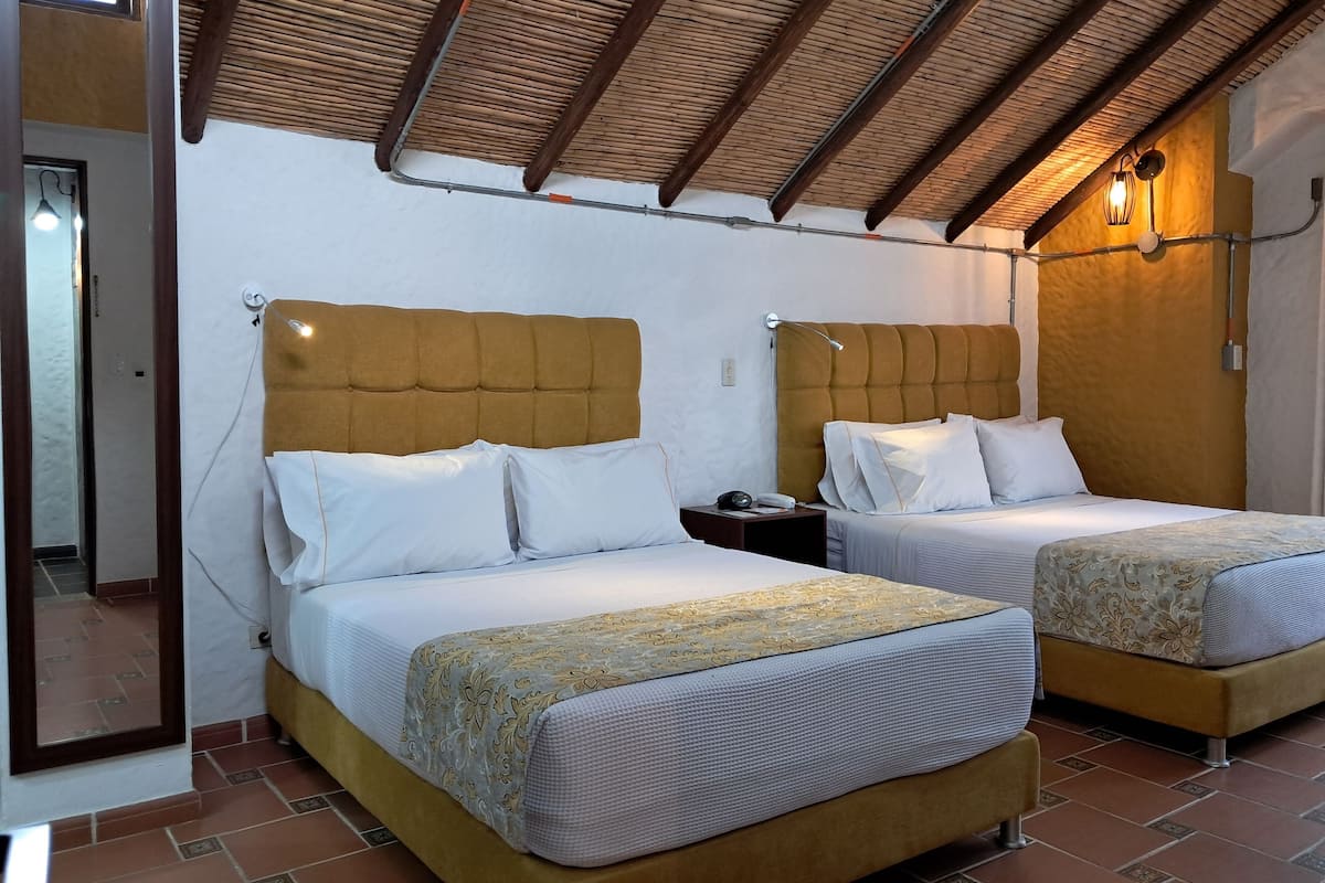Una habitación con dos camas, cada una con cabecera de madera, ropa de cama blanca y colcha estampada. Hay una mesita de noche con lámpara y teléfono en una de las camas. El suelo es de baldosas y el techo inclinado presenta vigas de madera.