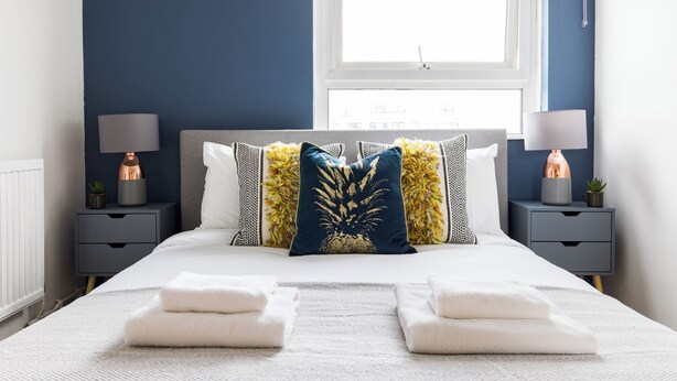Una cama bien hecha con sábanas blancas y dos toallas, una pared azul oscuro, una ventana con vistas y dos mesitas de noche con lámparas y plantas pequeñas.