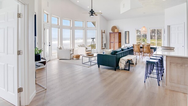 Ein geräumiger Wohnbereich mit großem Fenster zum Meer, ein dunkelgrünes Sofa, ein Essbereich mit Korbstühlen und eine Küche mit weißer Arbeitsplatte.