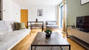 A modern living room with a sofa, a coffee table with a plant, a TV on a stand, and a dining area with a table and chairs. - The Dalston Apartments (London)