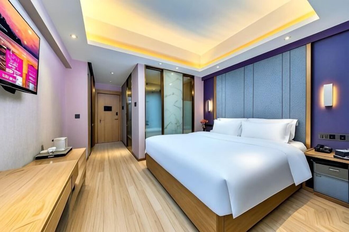 A modern hotel room with a large bed, a flat-screen TV, a desk with a chair, and a sliding glass door leading to a bathroom.
