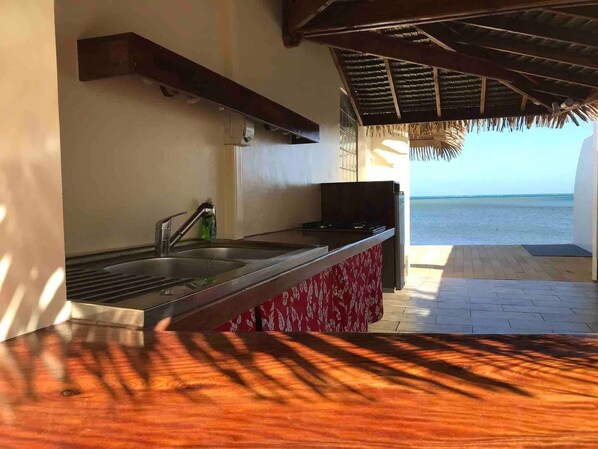 A kitchen with a view of the sea, a thatched roof, and a wooden counter. - Ocean Front Bungalow (Tumaraa)