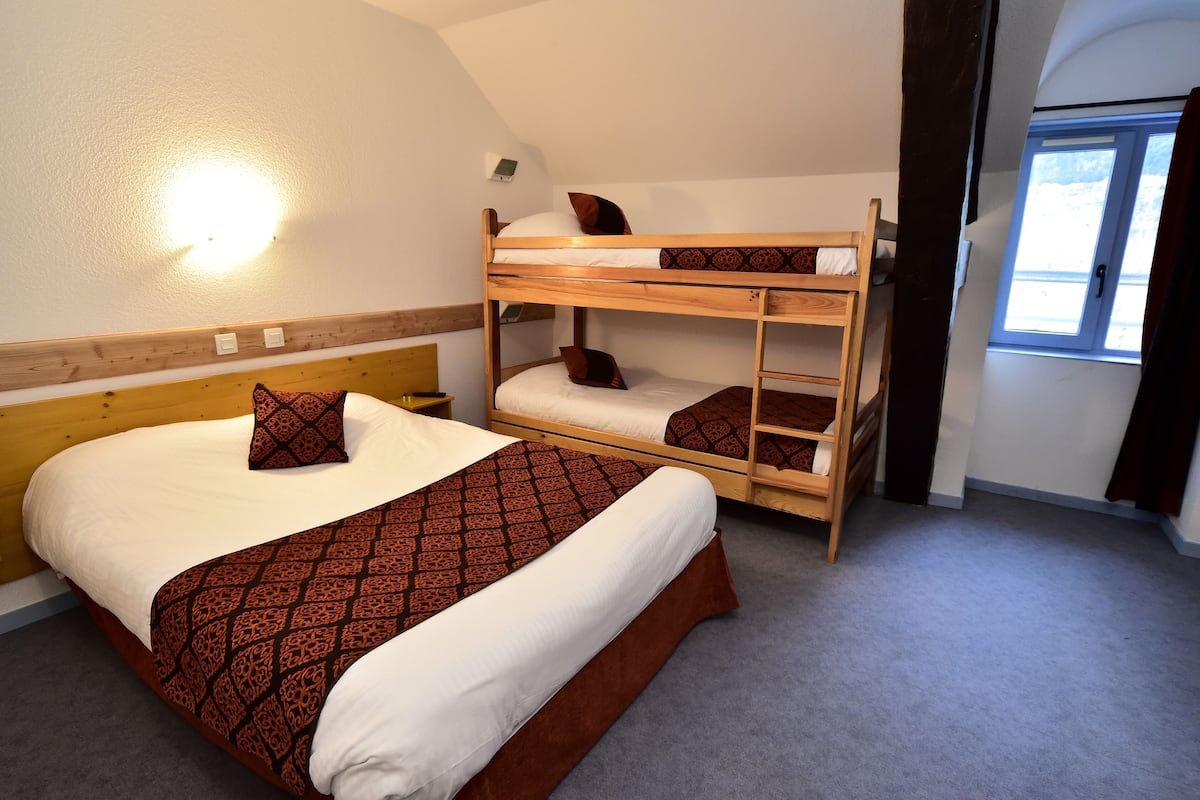 Une chambre avec un lit superposé doté d’une tête de lit en bois, un lit simple avec une literie à motifs et une fenêtre avec des rideaux.