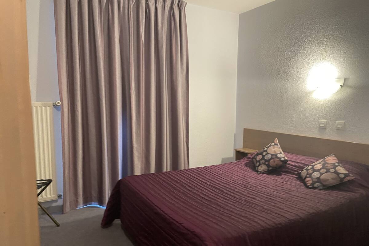 Une chambre d’hôtel avec un lit, des rideaux, une lampe fixée au mur et un radiateur.