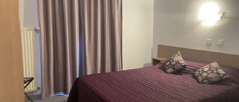 A hotel room with a bed, curtains, a wall-mounted light, and a radiator.
