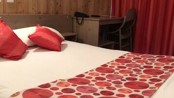 Et hotelværelse med en trævæg, et skrivebord, en stol og en seng med røde puder og et mønstret tæppe.