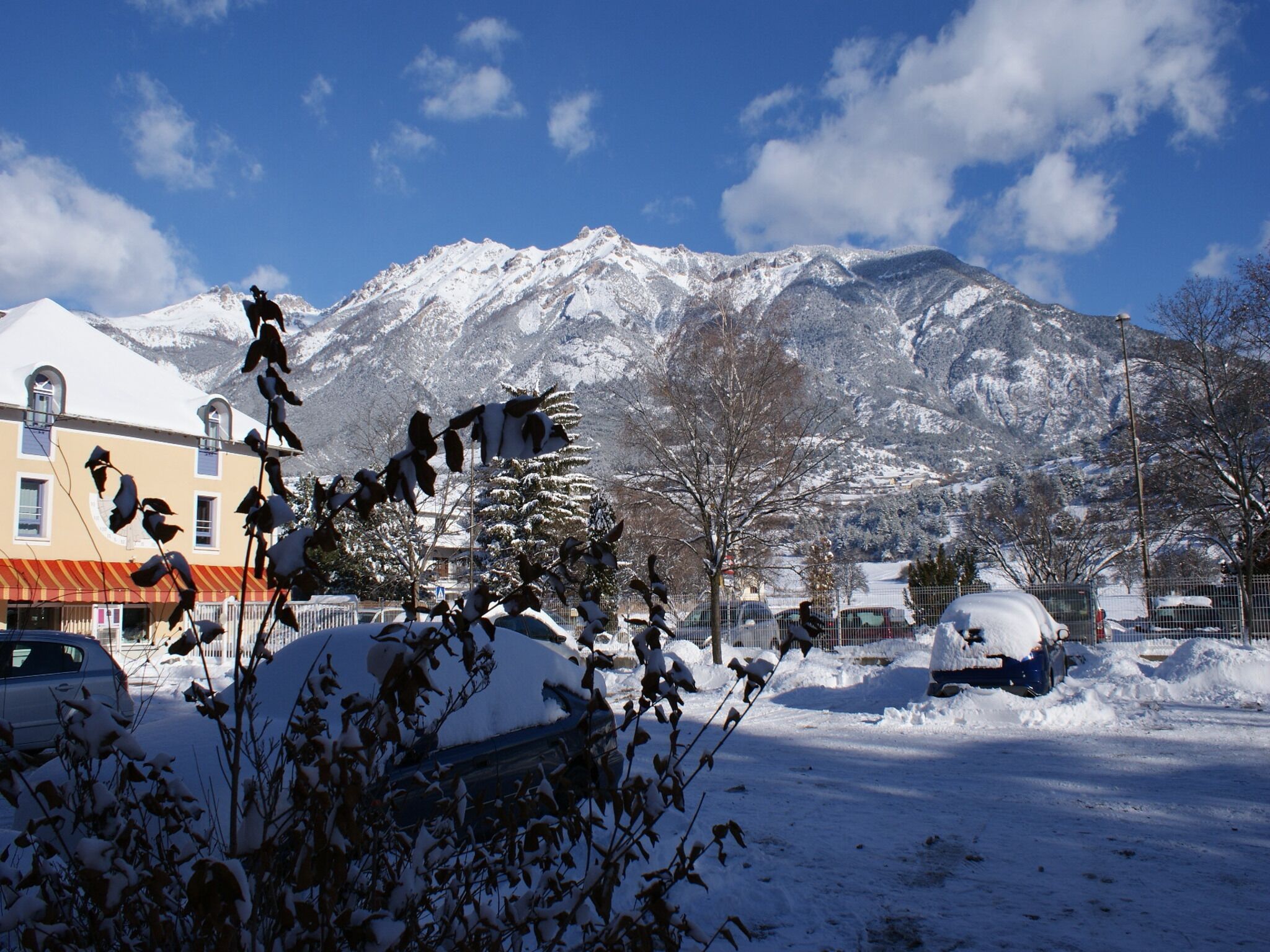 A snowy landscape with a mountain in the background, a building with a red roof, and parked cars.