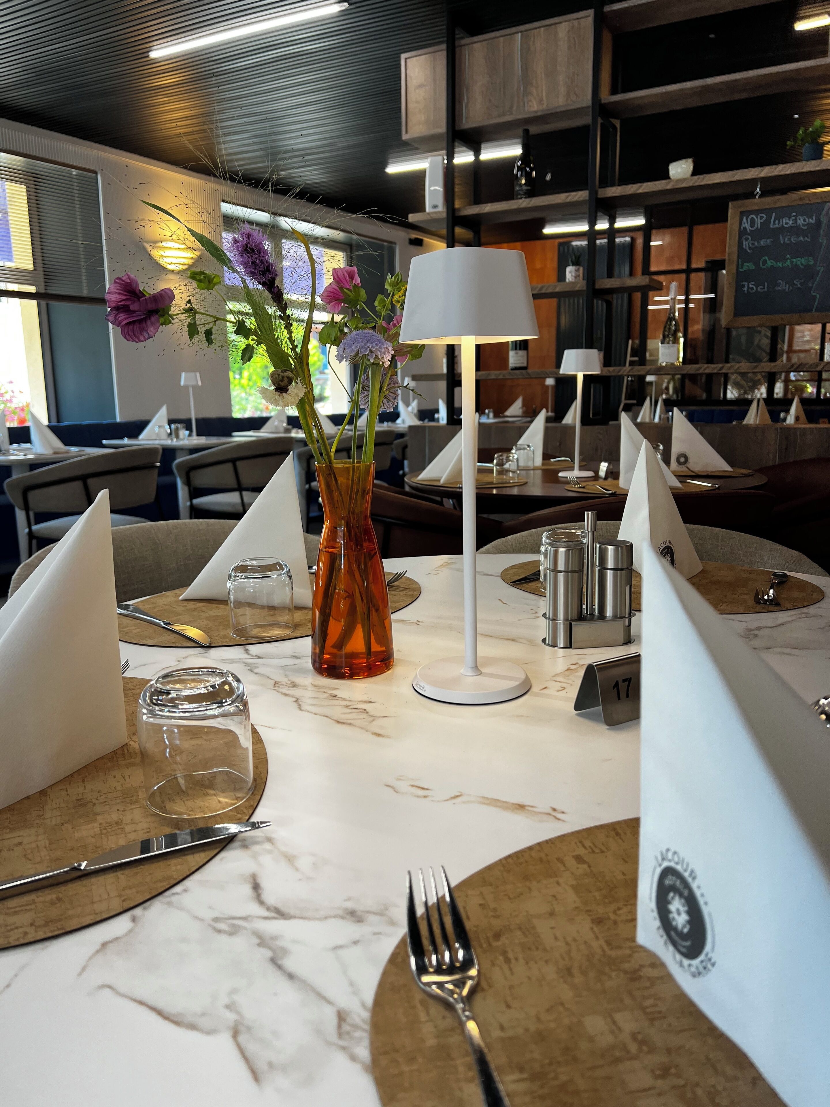 A restaurant setting with tables set for dining, featuring a vase with flowers, a lamp, and a menu board in the background.