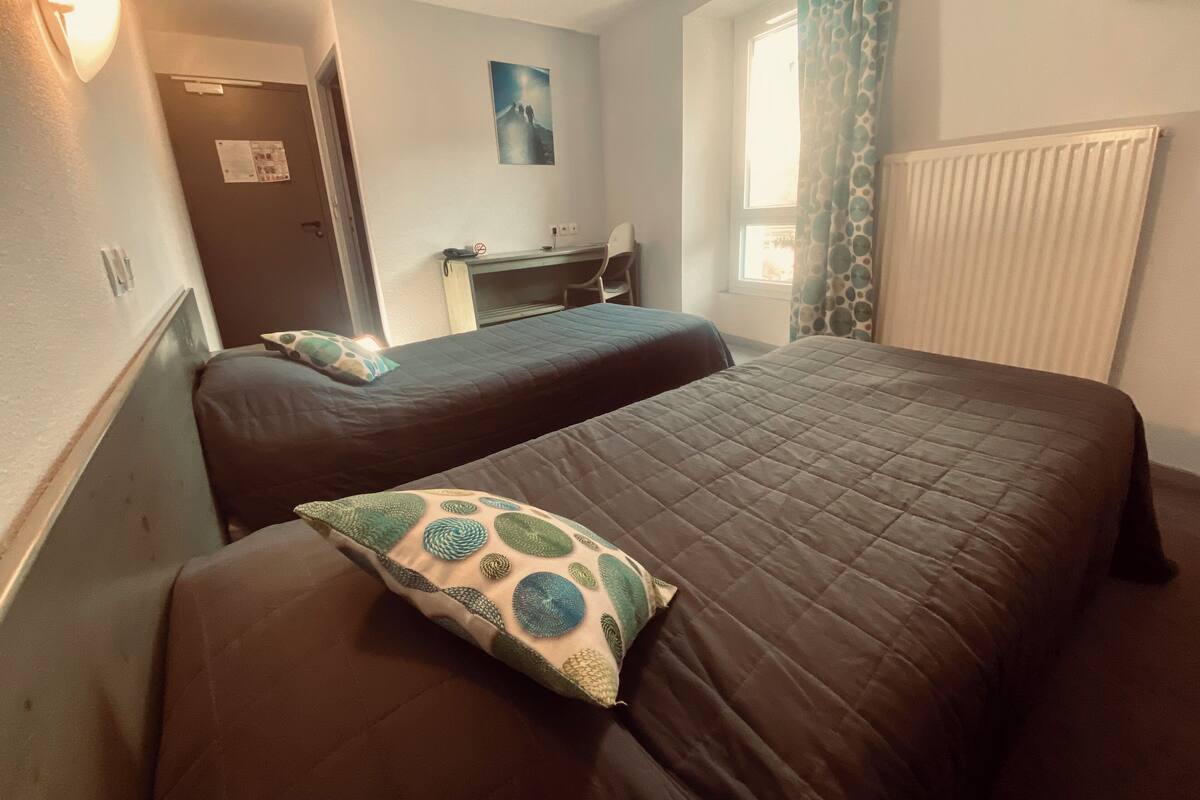 Une chambre d’hôtel avec deux lits, un bureau, une chaise, une fenêtre avec des rideaux et un radiateur.