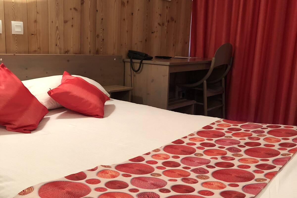 Une chambre d’hôtel avec un mur en bois, un bureau, une chaise et un lit agrémenté d’oreillers rouges et d’une couverture à motifs.