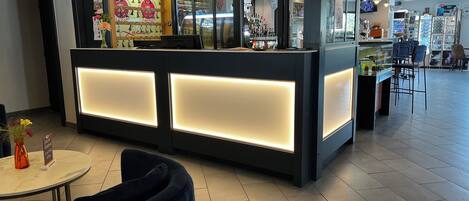 Un espace d’accueil au design moderne, comprenant un comptoir d’accueil doté de panneaux lumineux, un coin salon avec une table ronde et des chaises, et un bar visible à l’arrière-plan.