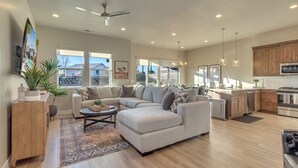 A spacious living room with a sectional sofa, a coffee table, and a rug. There is a kitchen area with wooden cabinets and stainless steel appliances.
