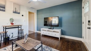 A living room with a dark wood floor, a white entertainment unit with a flat-screen TV, a ceiling fan, and a dining area with a table and bar stools. - 5min-Grant park|Fenced Yard|Parking|Fire Pit&BBQ (Atlanta)