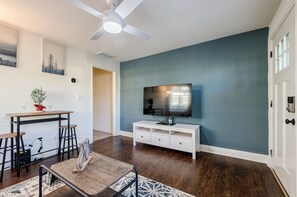 Un salon doté d’un parquet en bois foncé, d’un meuble de rangement blanc abritant un téléviseur à écran plat, d’un ventilateur de plafond et d’un coin repas avec une table et des tabourets de bar.