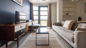 A modern living room with a dark blue accent wall, a flat-screen TV, a wooden cabinet, a glass-top coffee table, a beige sofa, and a large window with curtains. - Beautiful Canal-Side Apartment in Islington (London)