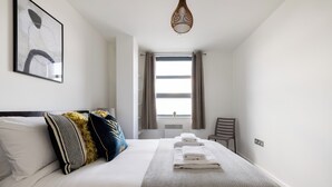 A neatly made bed with white linens and a patterned bedspread, a wall-mounted abstract painting, a wooden chair, and a window with curtains. - Beautiful Canal-Side Apartment in Islington (London)