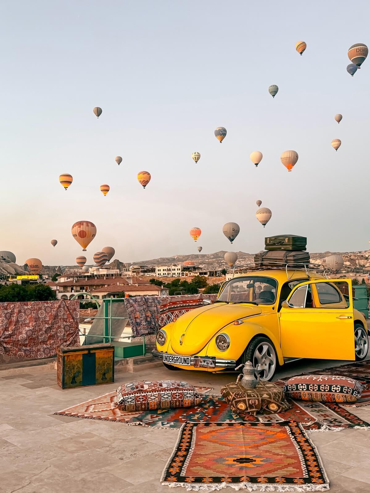 Un coche amarillo aparcado en una azotea con vistas a un paisaje urbano y globos aerostáticos en el cielo.