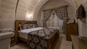A bedroom with a wooden bed, a stone wall, a stone bench, and a television on the wall. - Underground Cave Suites (Avanos)
