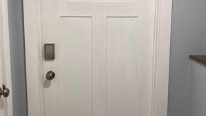 Une porte blanche dotée d’un clavier pour ouvrir la serrure et d’un trou de serrure, située dans un couloir aux murs de couleur claire et avec une étagère en bois sur le côté droit.