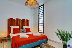 Een moderne slaapkamer met een houten hoofdeinde, een groot raam met horizontale jaloezieën, een rood-blauwe sprei en een pot met een varen.