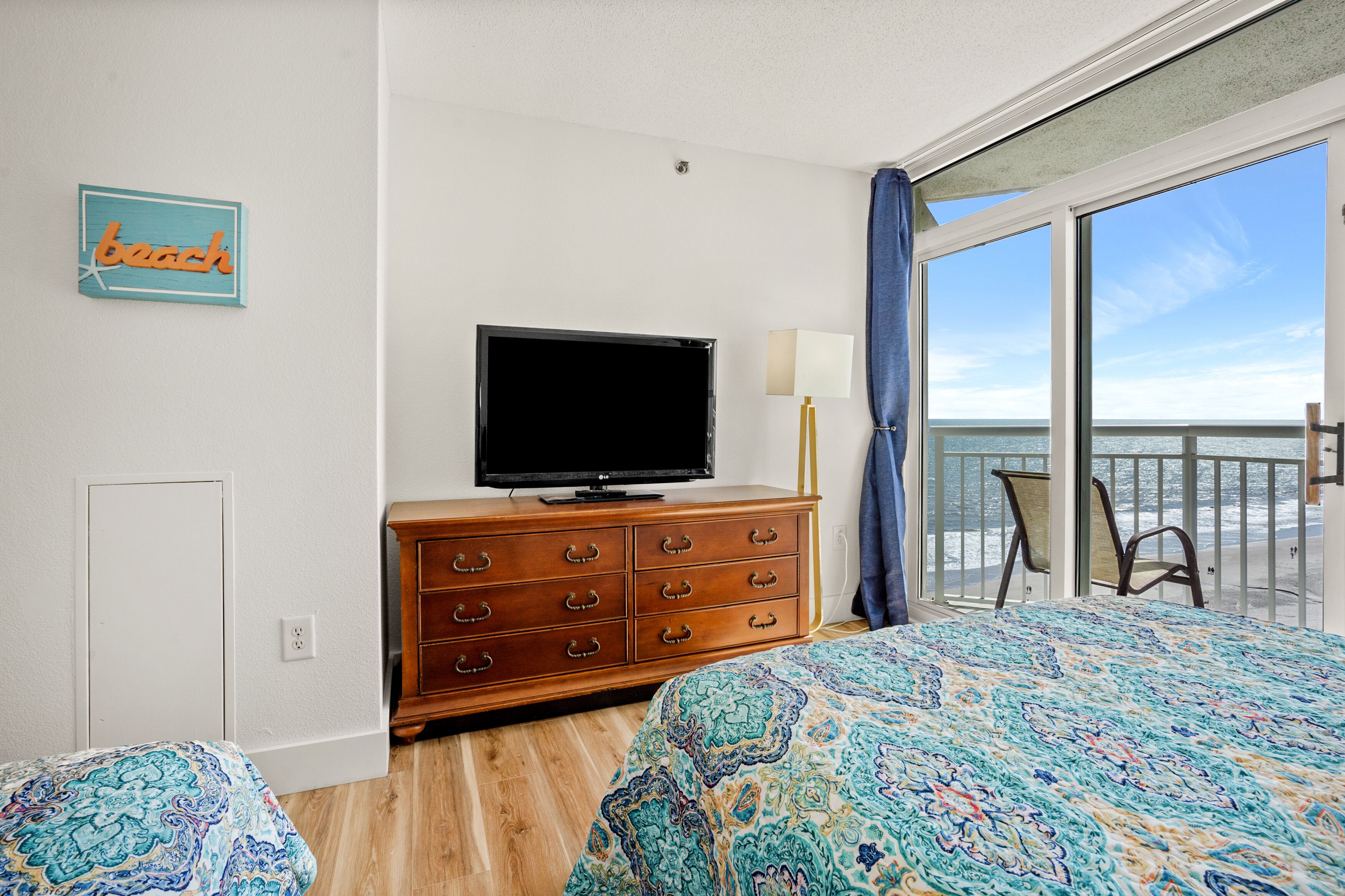 A bedroom with a large bed, a TV on a wooden dresser, a view of the sea through a balcony, and a 'beach' sign on the wall.