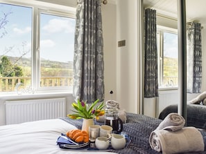 Ein ordentlich bezogenes Bett mit einem Tablett mit Frühstücksartikeln, darunter Croissants, Kaffee und Tee, in einem Zimmer mit großen Fenstern und Balkonblick.