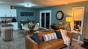 Ein Wohnzimmer mit braunem Ledersofa, einem Esstisch mit Stühlen, einer Küchenzeile und einer Wanduhr.