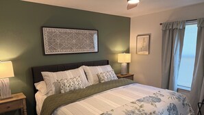 Ein Schlafzimmer mit einem Bett, zwei Nachttischen, einem Wanddekor, einem Fenster mit Vorhängen und einem Deckenventilator.