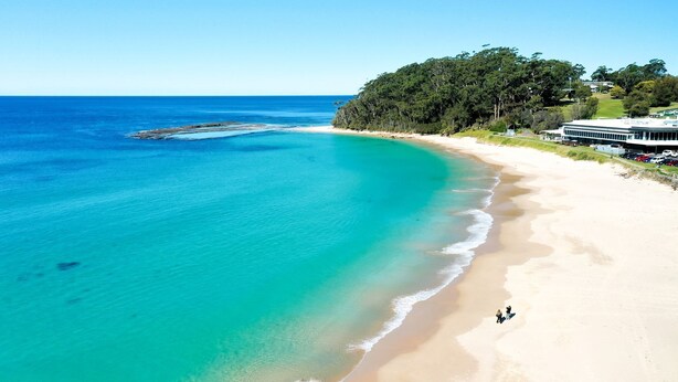 A beach with clear turquoise waters, a sandy shore, and a building with a large window overlooking the beach. - Breakers 4 - Bright spacious apartment in the heart of Mollymook (Mollymook)