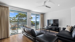 A modern living room with a large TV, leather sofas, and a glass door leading to an outdoor area. - Breakers 4 - Bright spacious apartment in the heart of Mollymook (Mollymook)