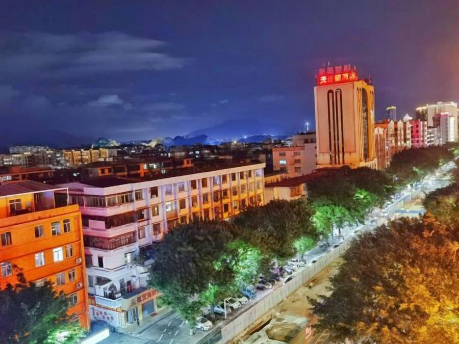 Un paisaje urbano nocturno con edificios iluminados, una torre destacada con señalización roja y una calle arbolada.