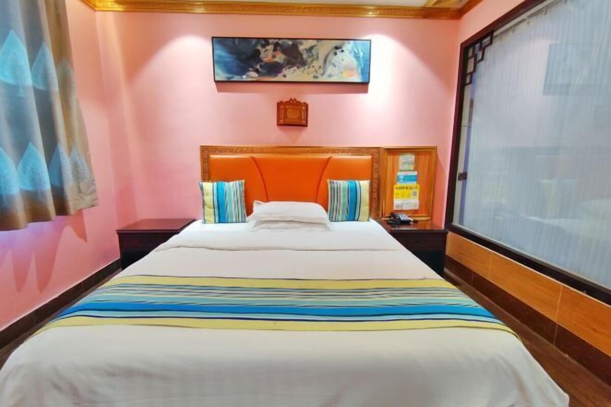 A hotel room with a large bed, a headboard with a decorative panel, a painting above the bed, and a window with a view.