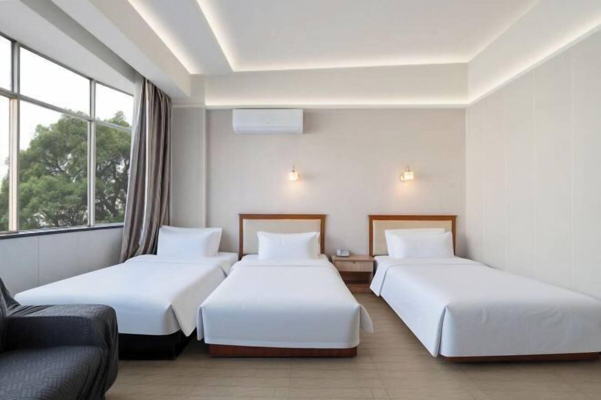 A hotel room with two single beds, a sofa, a window with curtains, and an air conditioning unit.