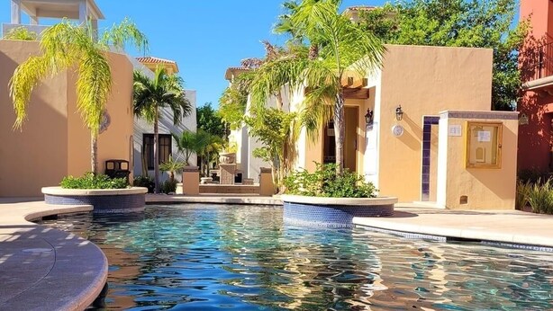 A pool area with a clear blue pool, surrounded by buildings with beige and white walls, palm trees, and a clear sky.