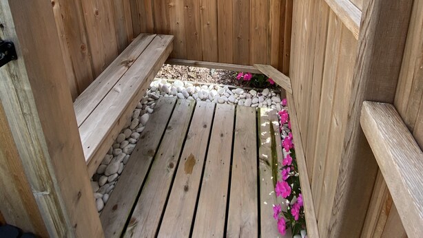 A wooden outdoor shower with a metal showerhead, wooden steps, and a pebble-lined floor. - Cape Escape Cottage (Yamouth)