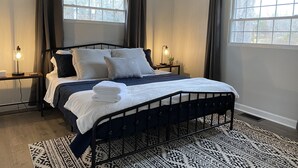 A neatly made bed with a white and navy blue bedspread, a folded white towel, and a patterned rug. There are two bedside tables with lamps, a ceiling fan, and a window with curtains.