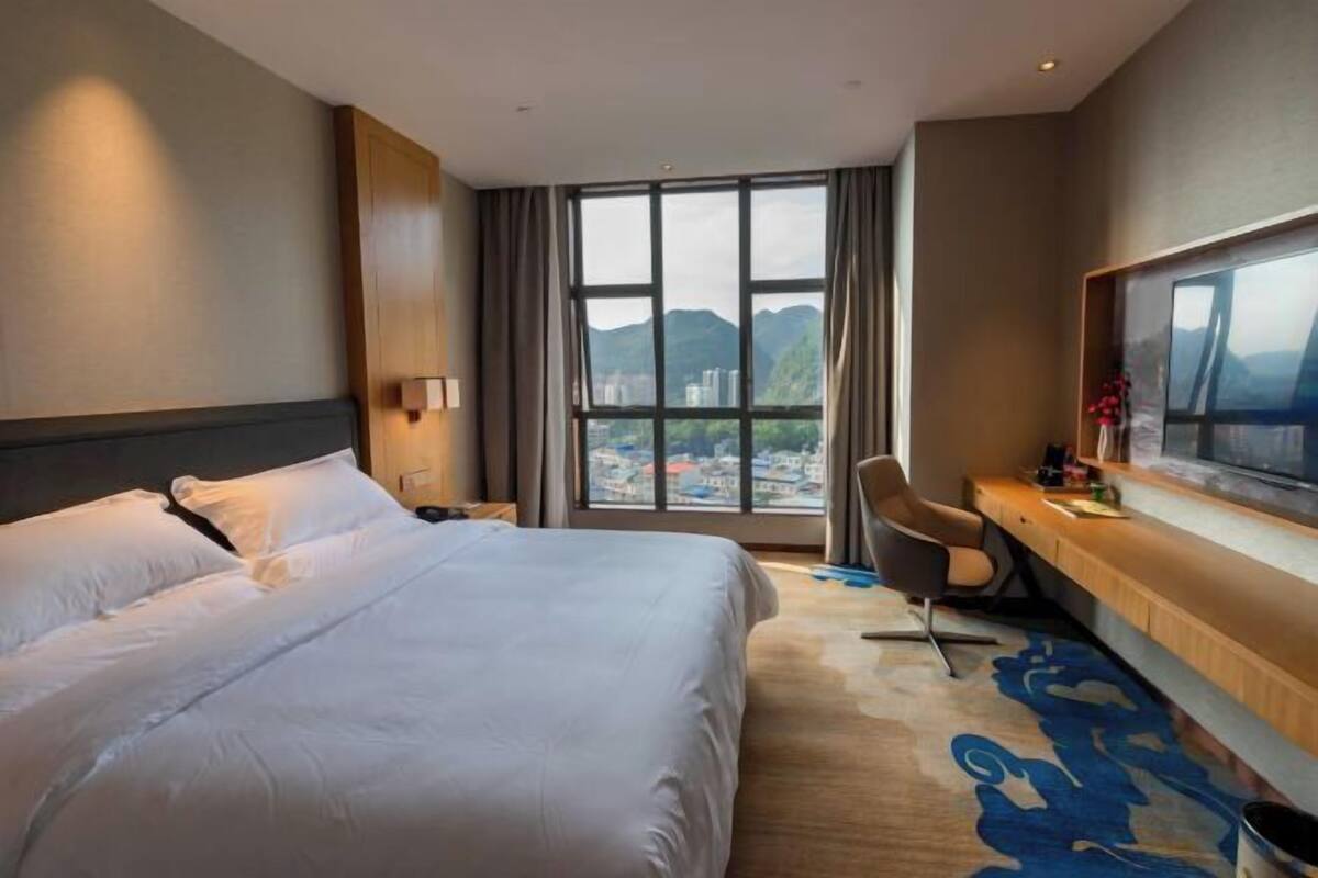 Habitación de hotel con una cama grande, un escritorio, una silla, un televisor y vistas a un paisaje urbano a través de amplios ventanales.