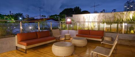 Una terraza en la azotea con mobiliario de mimbre, incluyendo sofás y sillones, y otomanas redondas.